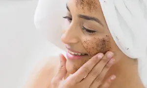 Kobieta z ręcznikiem na głowie nakłada na twarz peeling kawowy. Pokazuje, jak zrobić peeling dla promiennej cery.