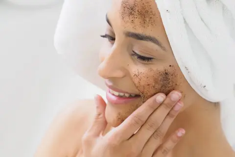 Kobieta z ręcznikiem na głowie nakłada na twarz peeling kawowy. Pokazuje, jak zrobić peeling dla promiennej cery.