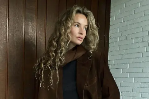 Kobieta z kręconymi blond włosami w brązowej marynarce. Kolor marynarki przypomina mi miód, taki **honey beige jaki to kolor** – ciepły i przytulny.