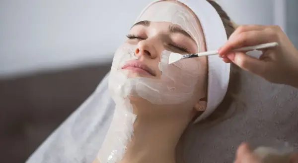 Kobieta z opaską na głowie otrzymuje peeling enzymatyczny nakładany pędzelkiem. Dowiedz się, jak stosować peeling enzymatyczny dla promiennej cery.