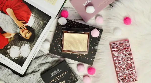Złoty rozświetlacz Anastasia Beverly Hills na czarnym opakowaniu z gwiazdami, obok różowego etui i magazynu.