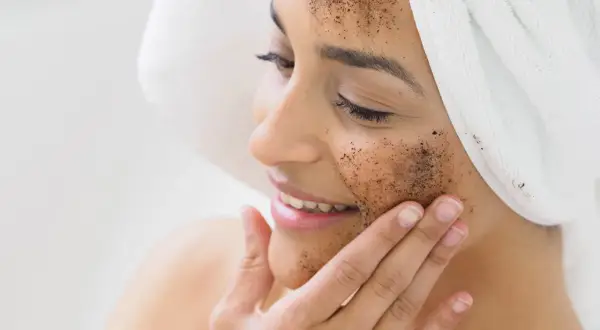 Kobieta z ręcznikiem na głowie nakłada na twarz peeling kawowy. Pokazuje, jak zrobić peeling dla promiennej cery.