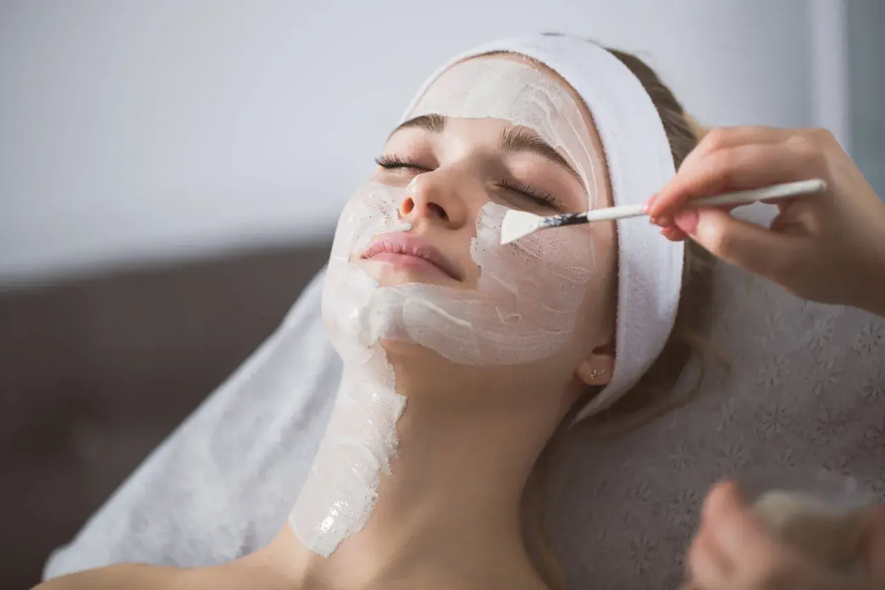 Kobieta z opaską na głowie otrzymuje peeling enzymatyczny nakładany pędzelkiem. Dowiedz się, jak stosować peeling enzymatyczny dla promiennej cery.