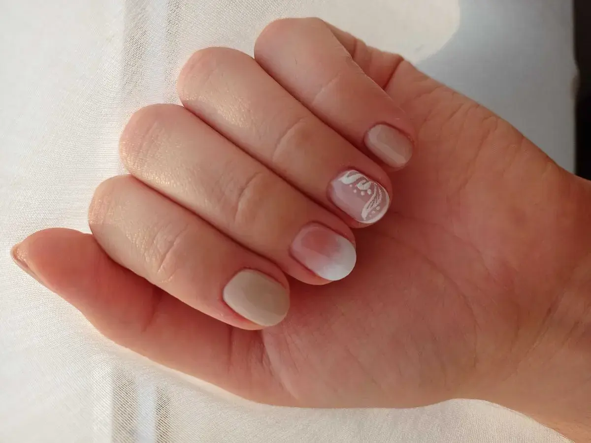 Delikatne rodzaje manicure: beżowe paznokcie z subtelnym zdobieniem białym wzorem na jednym paznokciu.