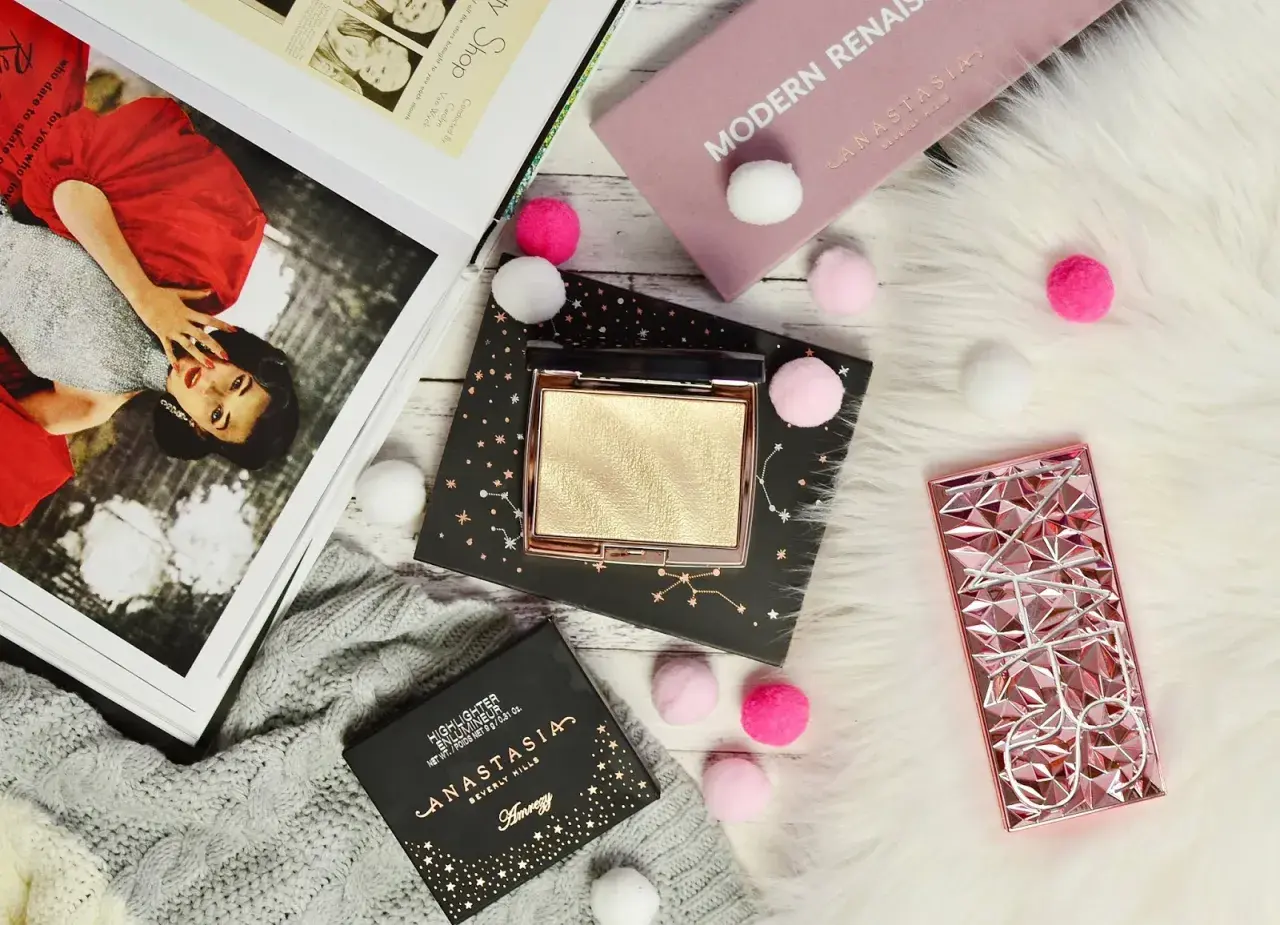 Złoty rozświetlacz Anastasia Beverly Hills na czarnym opakowaniu z gwiazdami, obok różowego etui i magazynu.