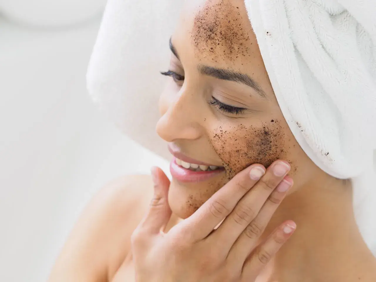 Kobieta z ręcznikiem na głowie nakłada na twarz peeling kawowy. Pokazuje, jak zrobić peeling dla promiennej cery.