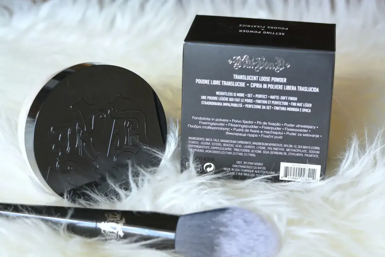 Czarny, błyszczący puder Kat Von D z wytłoczonym logo, leżący obok pudełka i pędzla na białym futerku.