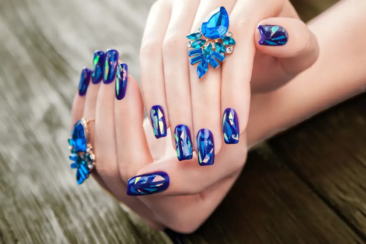 Dłonie z paznokciami w stylu glass effect nails, ozdobione niebieskimi pierścionkami z kamieniami.